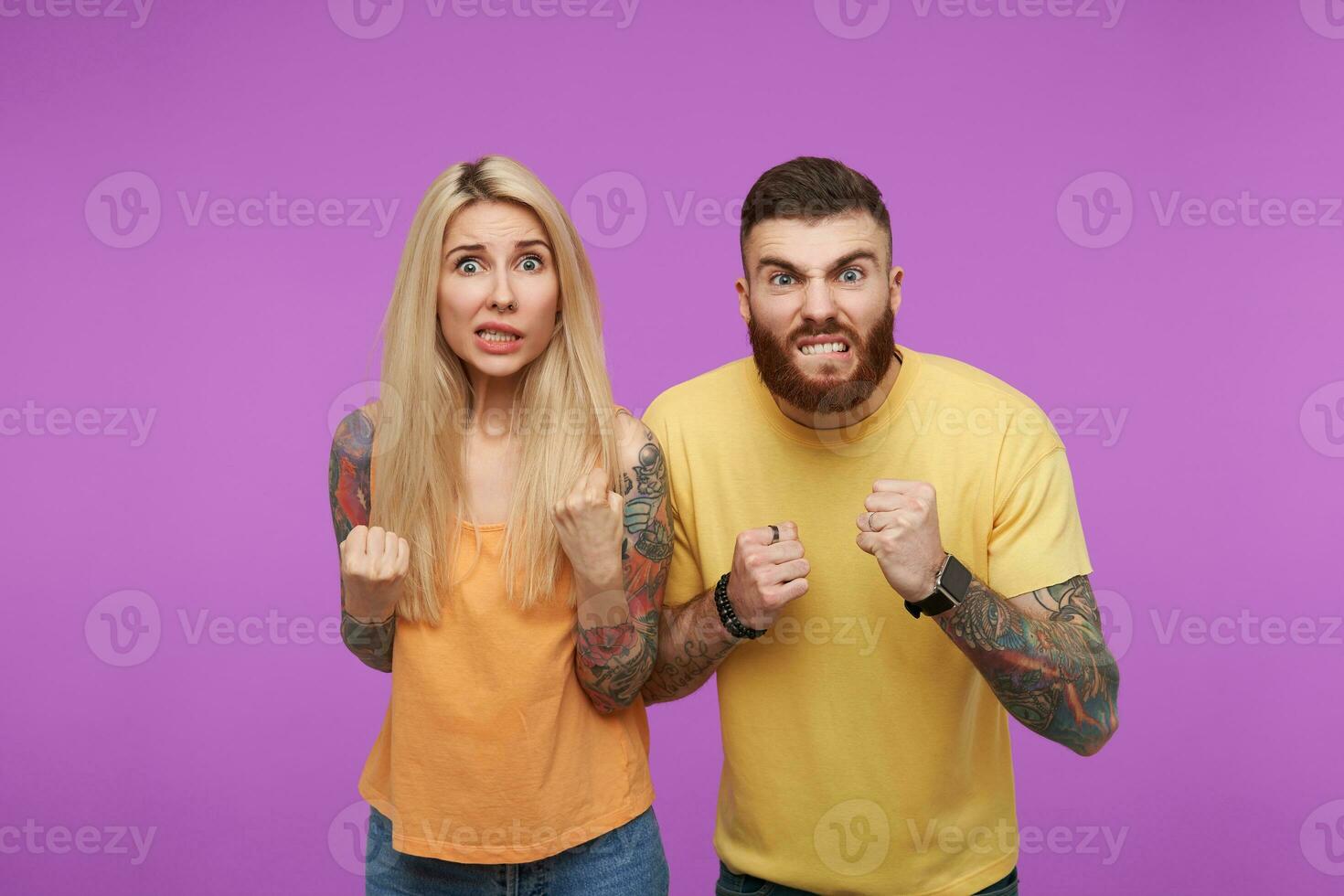 Horizontal photo of young excited tattooed couple folding fists with raised hands and showing teeth while grimacing faces, standing over purple background