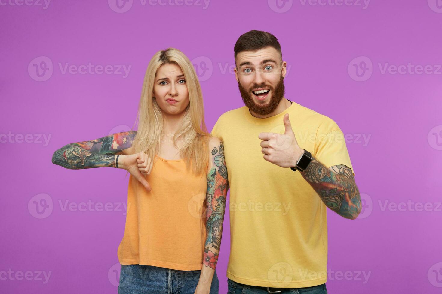 Horizontal shot of young pretty tattooed couple in casual wear keeping their hands raised while gesturing and expressing different emotions, isolated over purple background photo