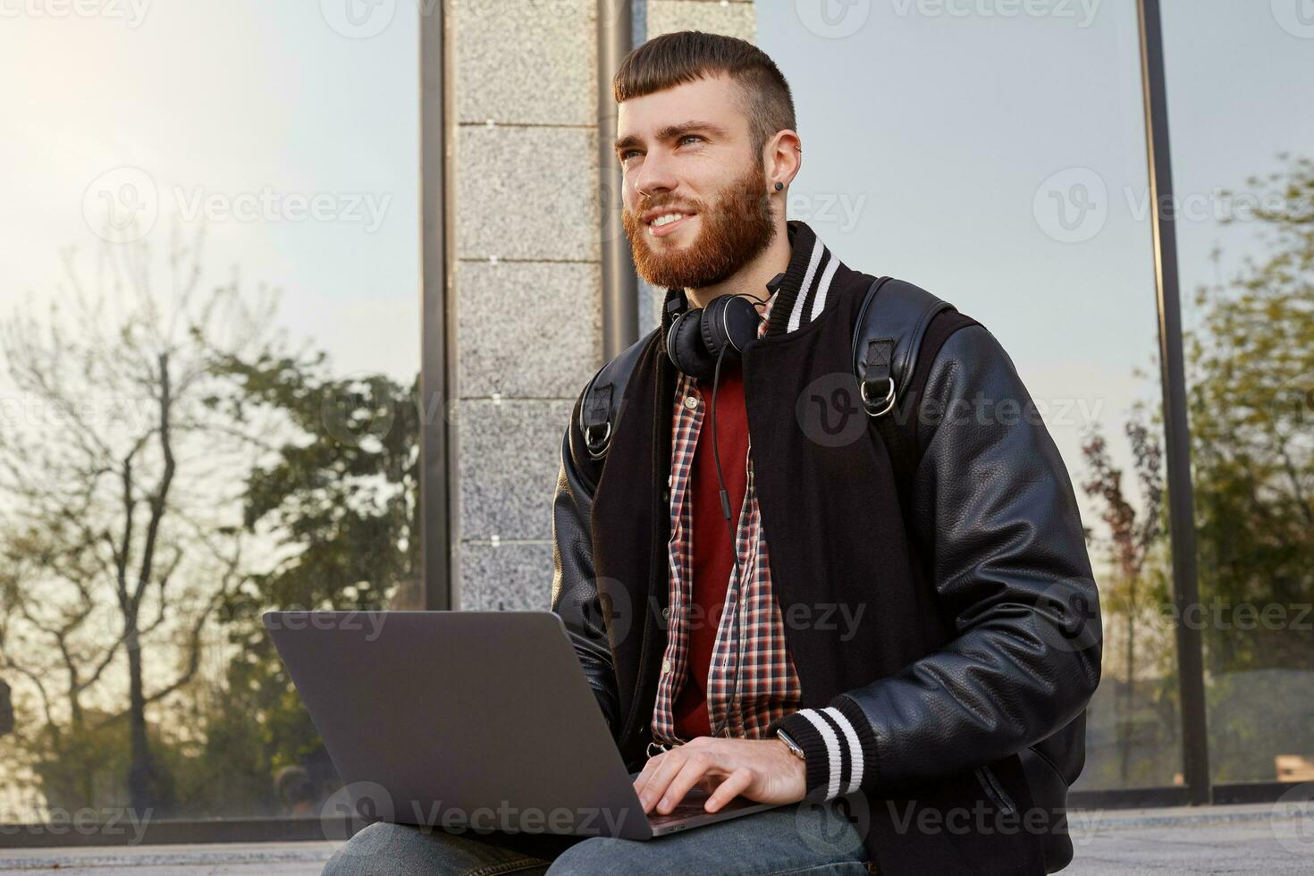 Outdoor shot of handsome red bearded young guy, sitting on the street putting the laptop on lap, looking into the distance and planning a trip to the mountains. photo