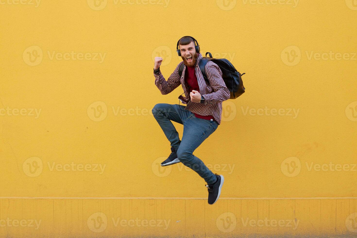 Smiling young attractive ginger bearded man jumping,happy success, wearing in basic clothes with backpack. Looking at the camera over a yellow wall with copy space. photo