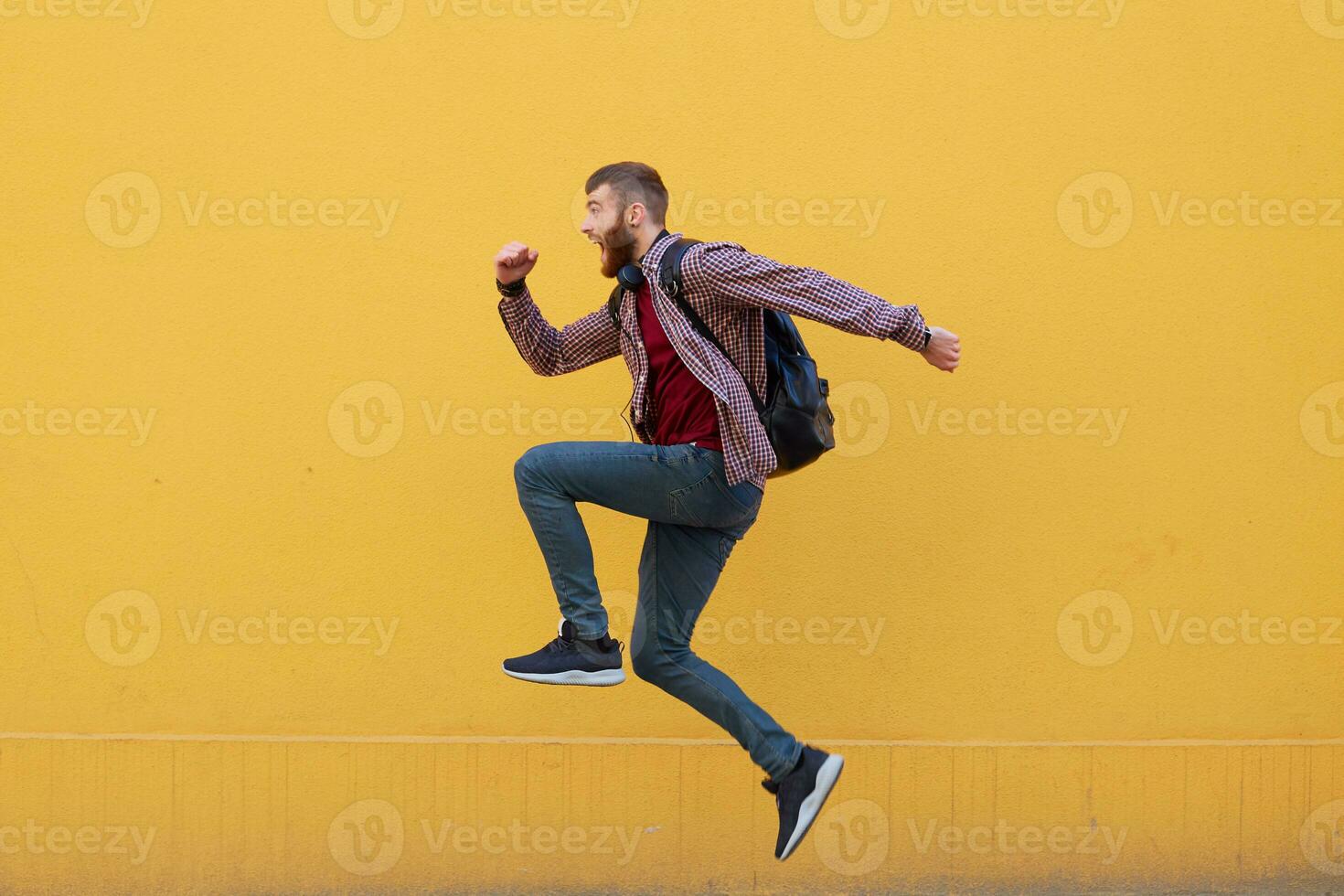 Young attractive ginger bearded man jumping, very fast running, hurry to the store for sale, wearing in basic clothes with backpack. Isolated over yellow wall with copy space. photo