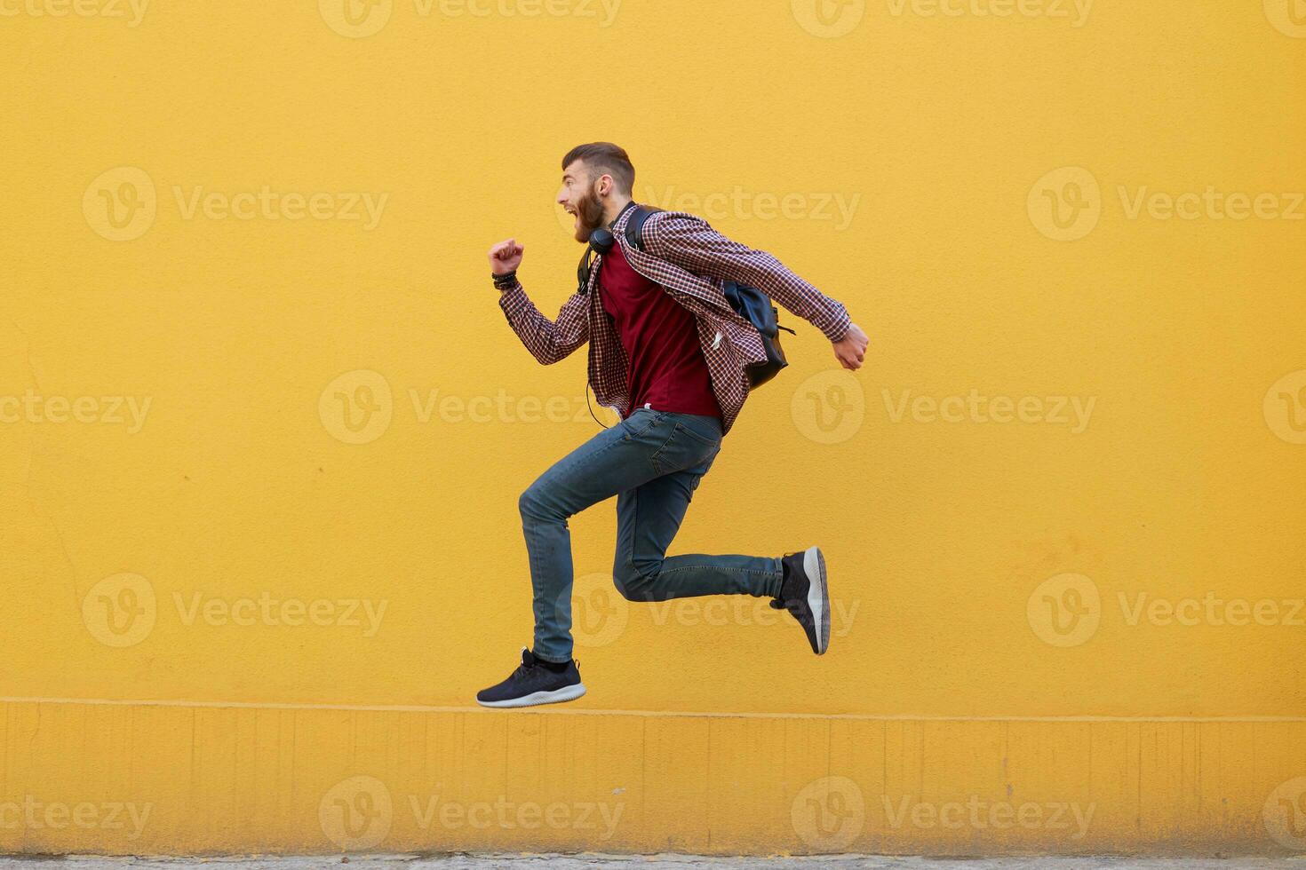 Young attractive ginger bearded man jumping, very fast running, running away from hateful work, wearing in basic clothes with backpack. Isolated over yellow wall with copy space. photo