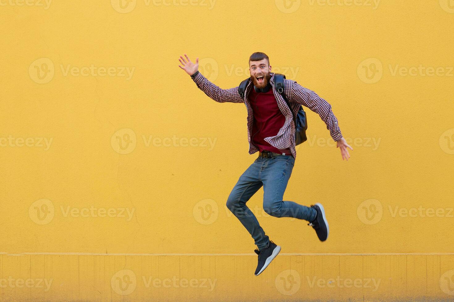 Happy young attractive ginger bearded man jumping and waving his hand, wearing in basic clothes with backpack. Looking at the camera, smiling with wide open mouth, over a yellow wall with copy space. photo