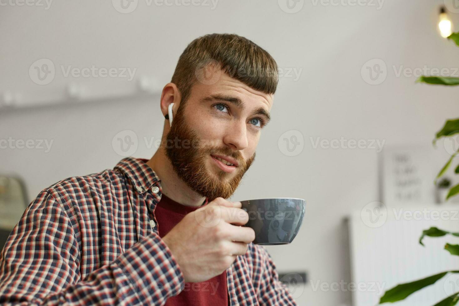 retrato de contento joven atractivo jengibre barbado hombre participación un gris café taza, soñando mira arriba y disfruta café, vistiendo en básico ropa. foto