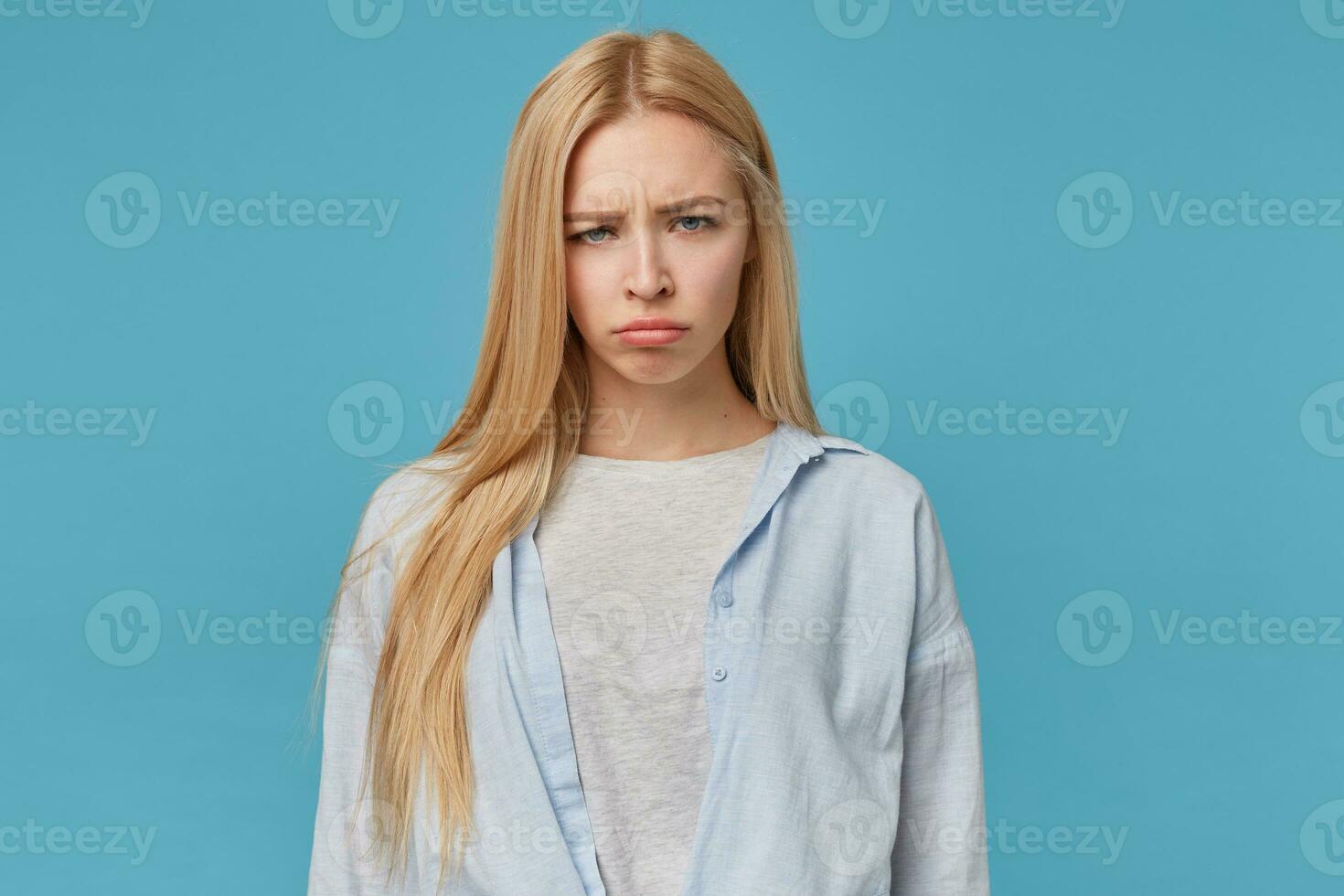 Close-up of offended blonde long haired female wearing blue shirt and grey t-shirt, looking at camera with folded lips and frowning, posing over blue background with hands along body photo