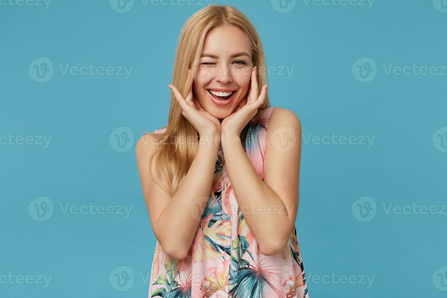 Horizontal photo of positive blonde long haired lady wearing flowered romantic dress, standing over blue background with charming smile, winking at camera and leaning head on raised palms