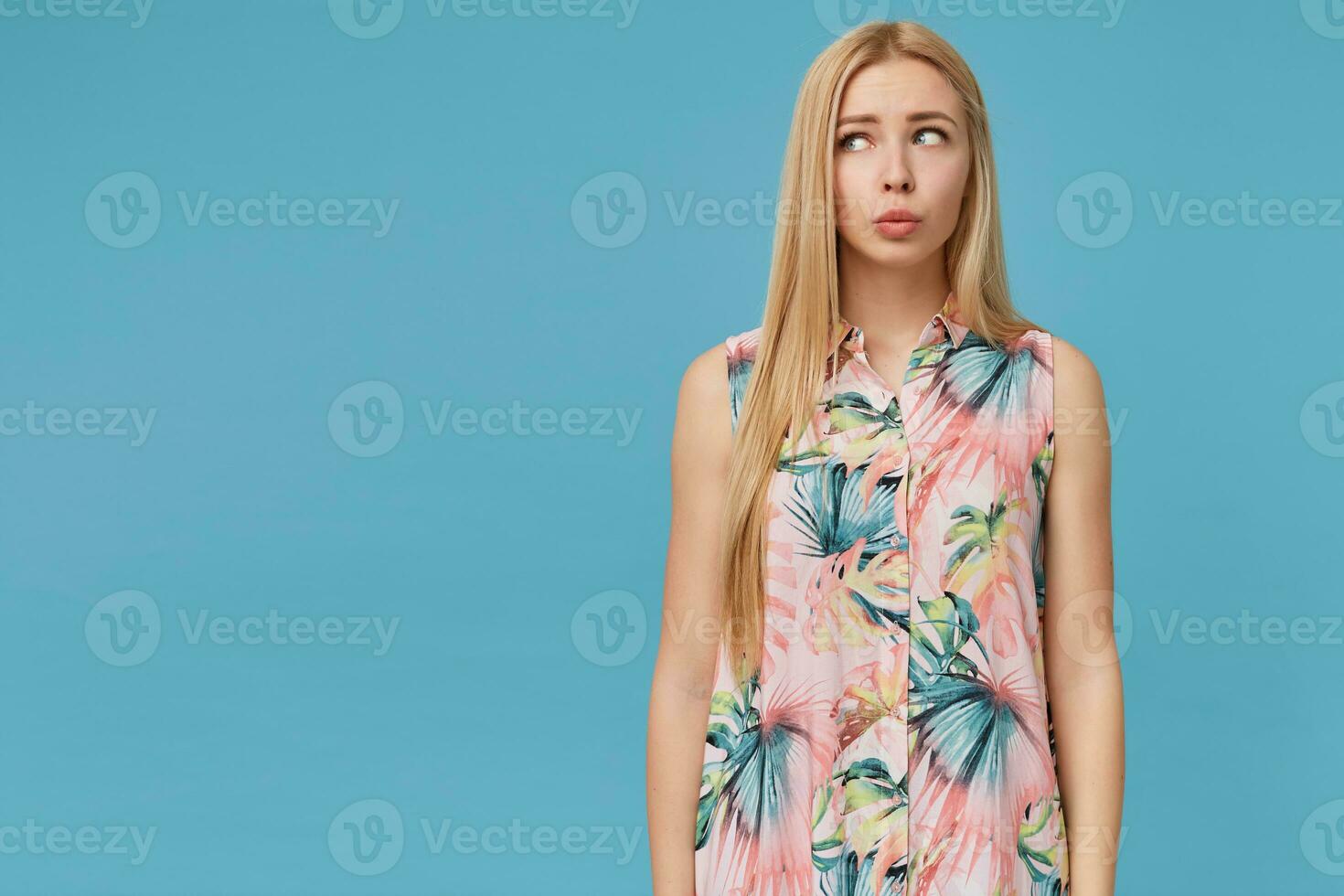 Studio photo of confused young blonde long haired lady wearing flowered shirt, posing over blue background with hands down, looking aside with pursed lips