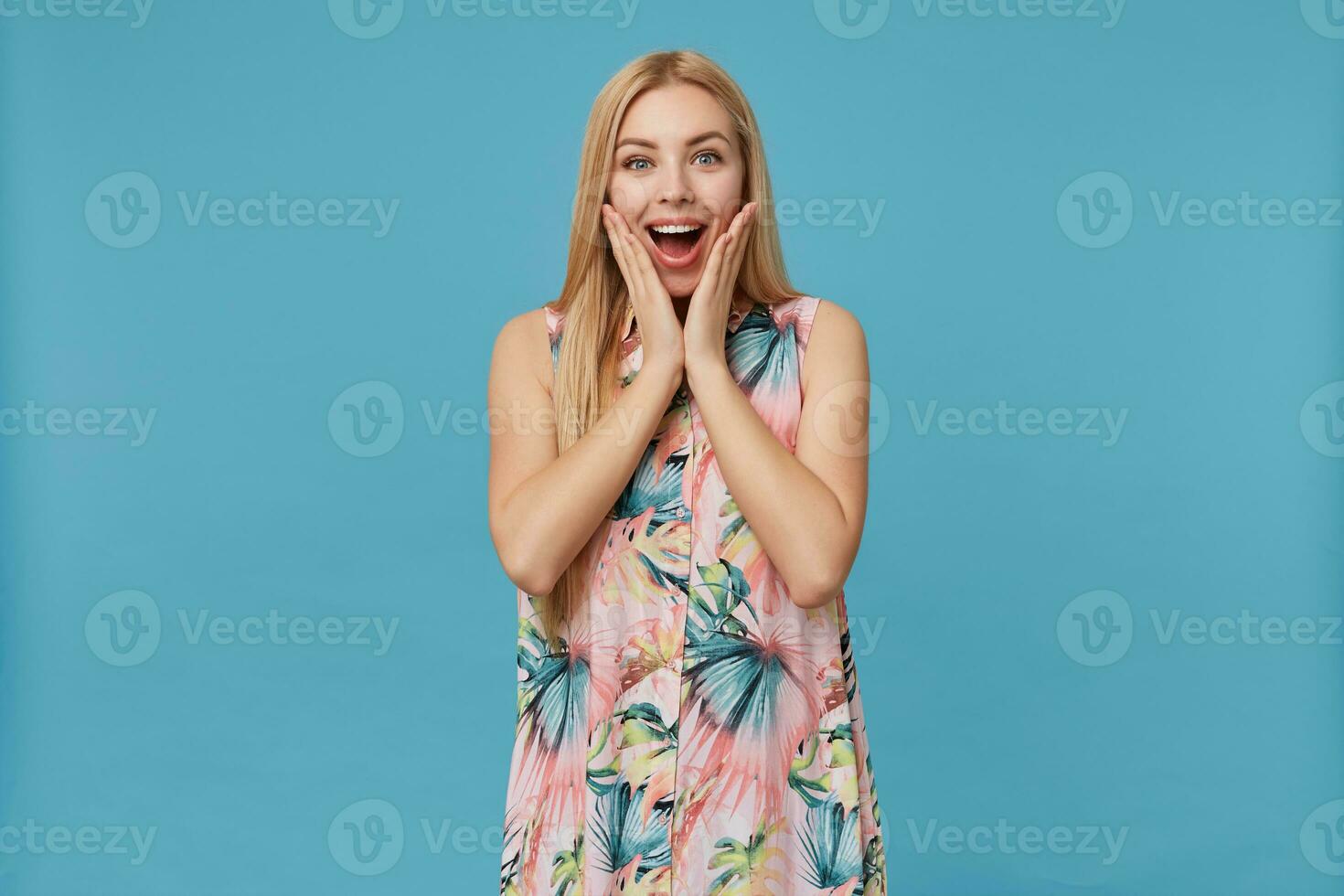 Studio photo of surpriesed young long haired blonde lady in flowered dress standing over blue background, holding hands on her cheeks and looking at camera surprisedly