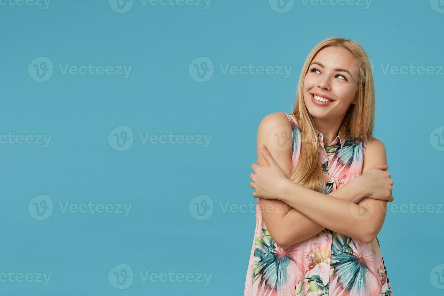 Lovely young female with blonde long hair wearing romantic dress with floral print, keeping hands crossed on her chest and looking aside with charming smile, isolated over blue background photo