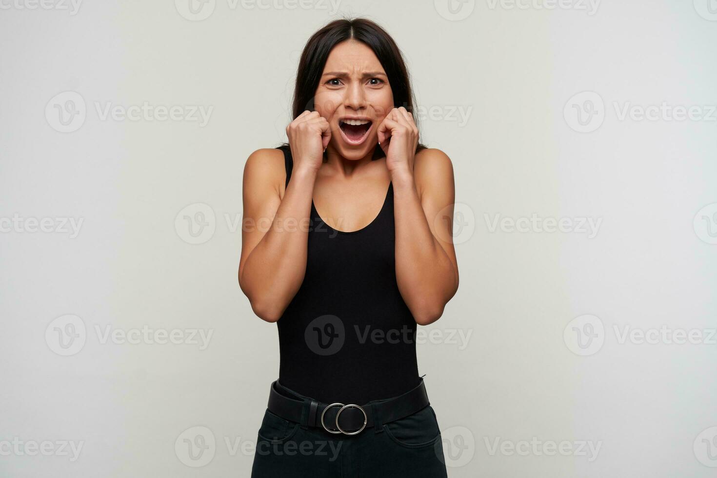 Frightened young dark haired woman with casual makeup raising hands to her face while looking scaredly at camera, frowning face with wide mouth opened while posing over white background photo