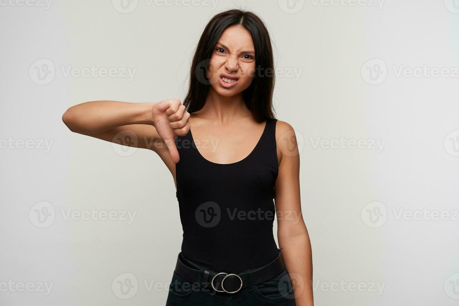 Displeased young brunette brown-eyed pretty woman showing down with thumb while looking at camera with pout and frowning her face, dressed in casual clothes while posing over white background photo