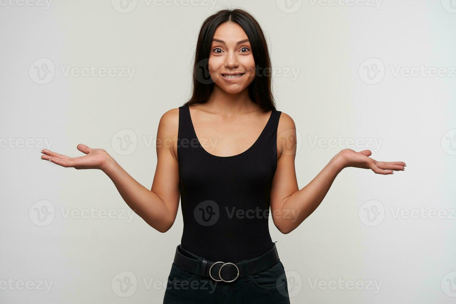 Cheerful young pretty brunette lady with casual hairstyle looking excitedly to camera and biting underlip, shrugging with opened arms while standing over white background photo