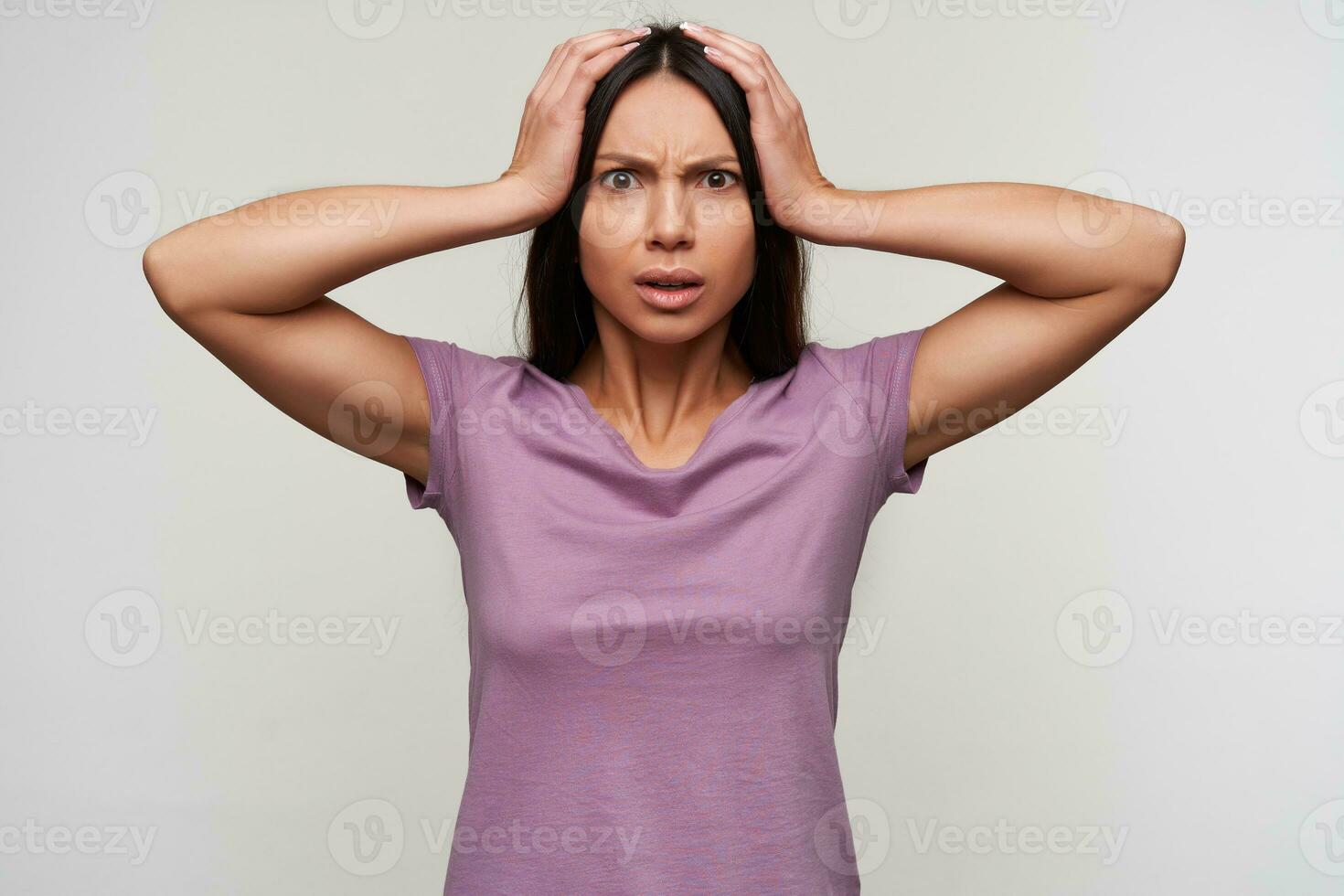 Bewildered young pretty brown-eyed brunette lady clutching her head and looking confusedly at camera, frowning her face while posing over white background in purple t-shirt photo