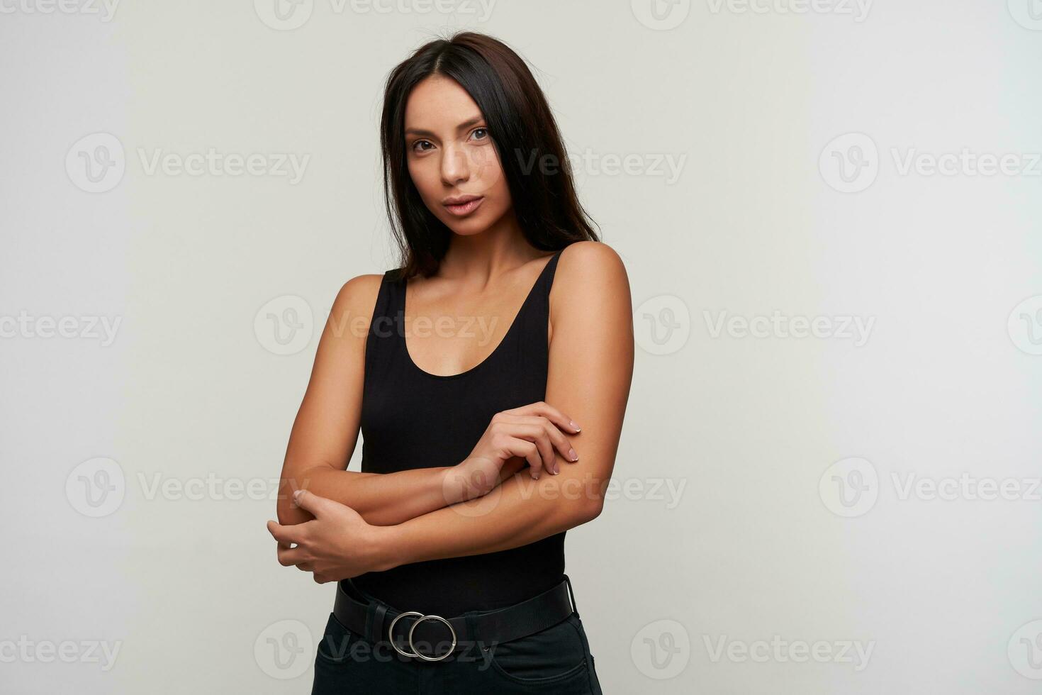 Charming young brown-eyed brunette lady folding her hands on chest and looking at camera with calm face, dressed in casual black clothes while posing over white background photo