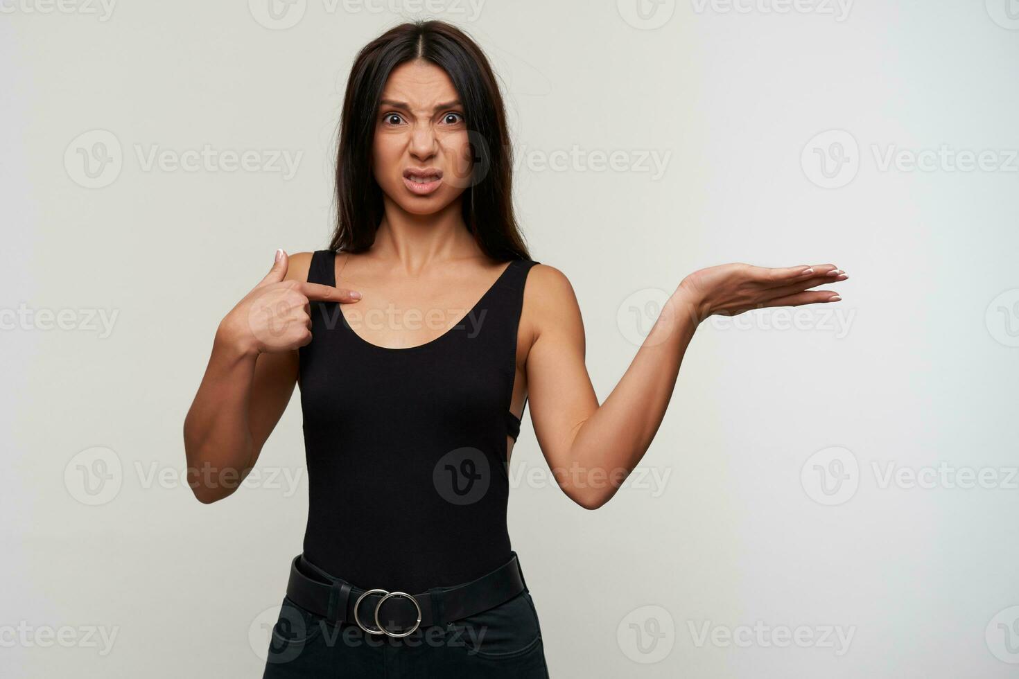 Studio shot of indignant young dark haired woman in casual black clothes pointing on herself with forefinger and frowning discontentedly her facce, isolated over white background photo