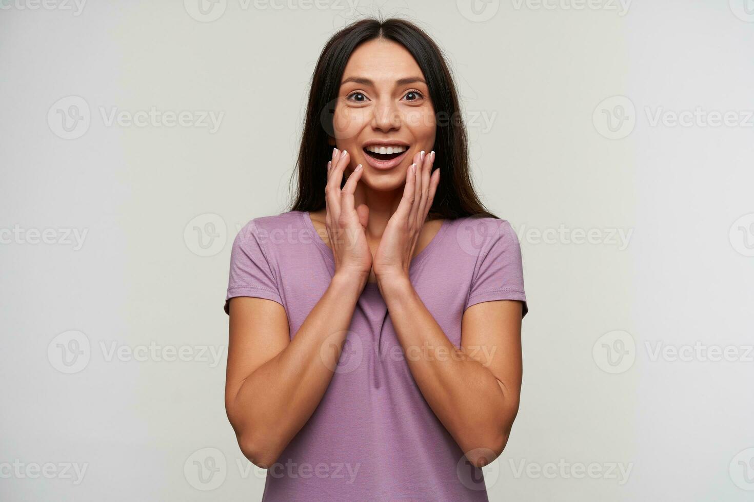 Surprised young attractive brunette lady with casual hairstyle looking at camera with wide eyes and mouth opened, keeping hands on her face while standing over white background photo