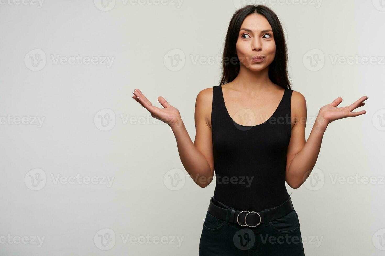 Suprised young attractive long haired brunette female rounding eyes while looking positively aside and raising palms up, dressed in casual black clothes while posing over white background photo