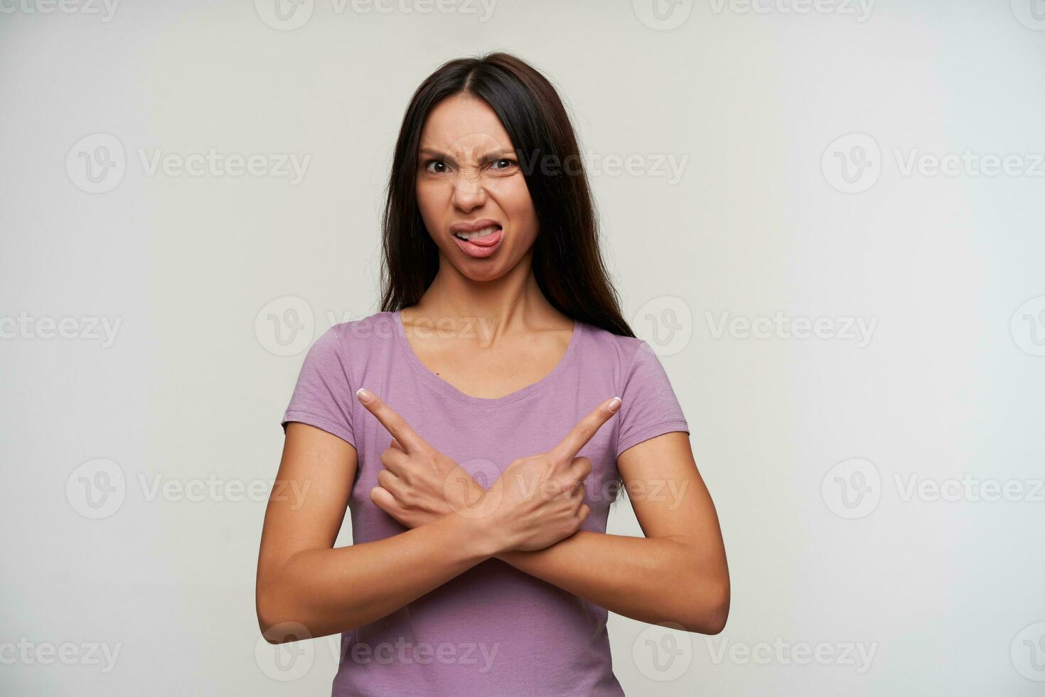 Dissatisfied young dark haired woman in purple t-shirt frowning her face and showing tongue at camera, pointing with forefingers in different directions, standing against white background photo