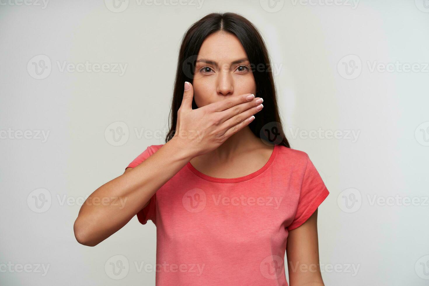 interior foto de hermosa joven de ojos marrones morena hembra vestido en rosado camiseta acuerdo palma en su boca mientras mirando a cámara, aislado terminado blanco antecedentes