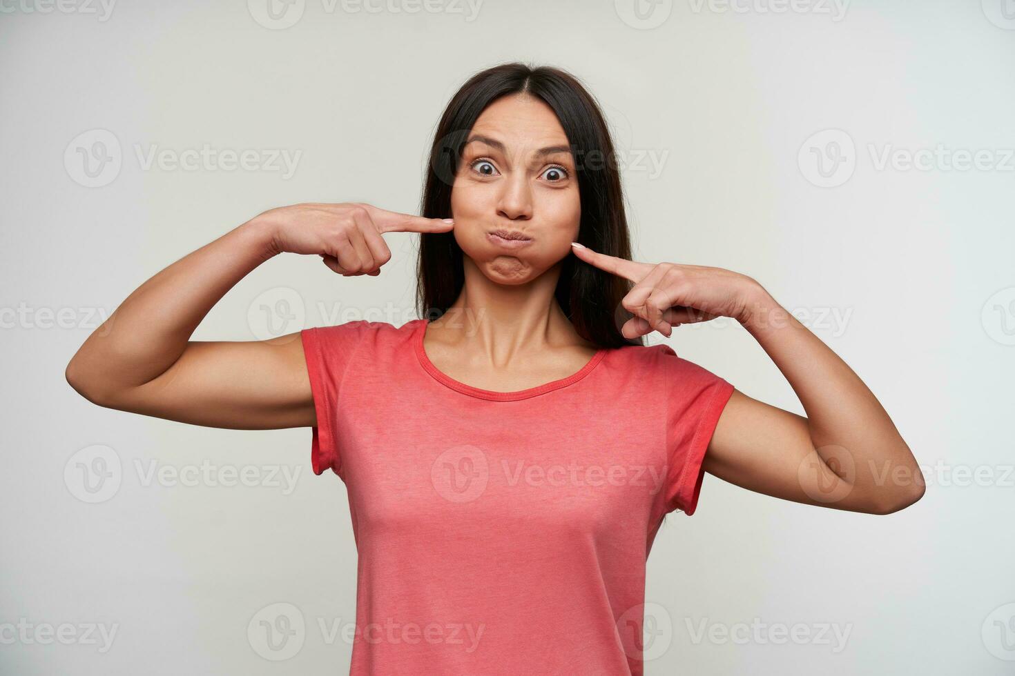 Funny shot of young brown-eyed brunette woman with natural makeup keeping raised forefingers on her pouting cheeks, fooling while standing over white background photo