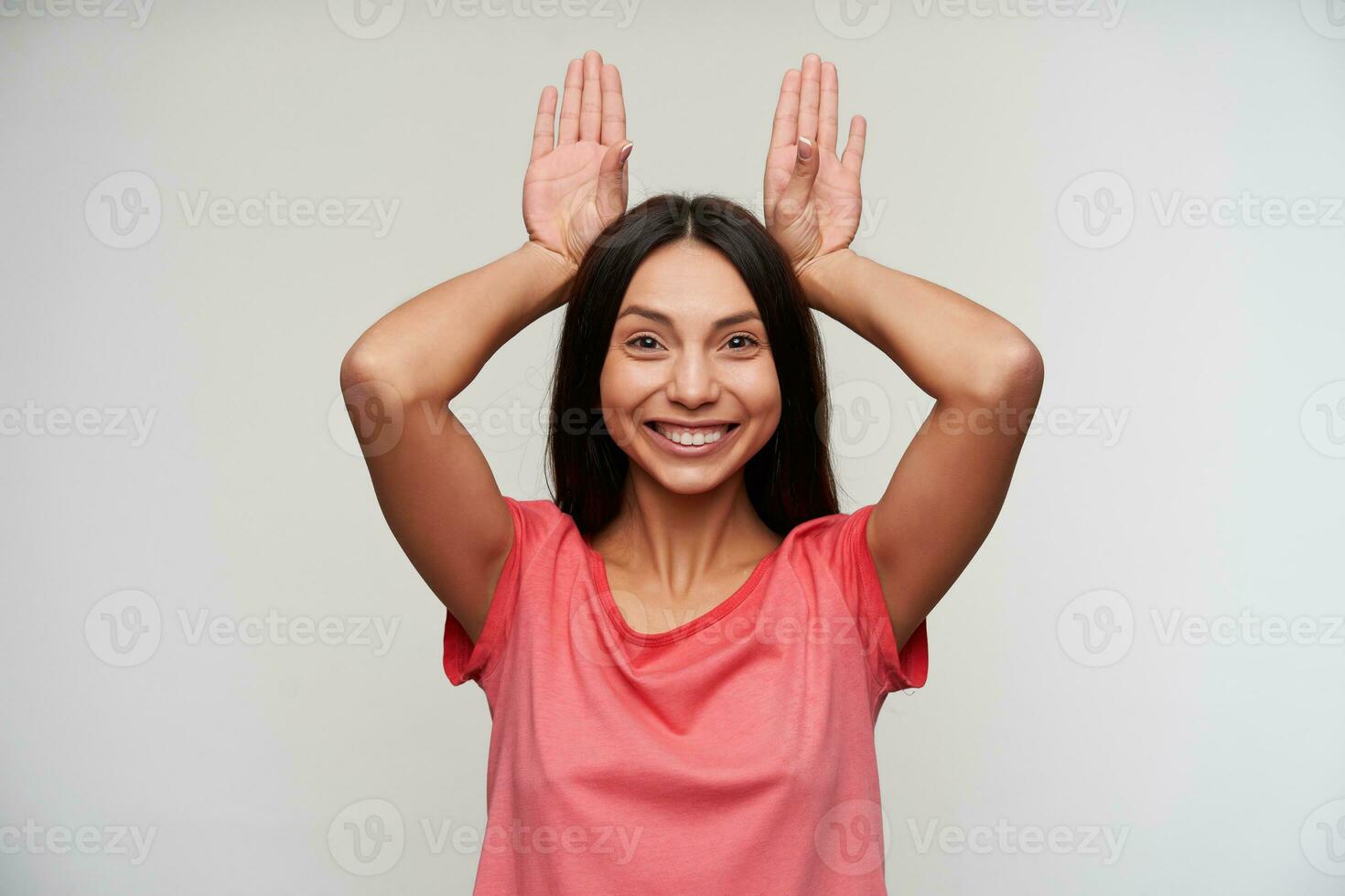 Indoor shot of positive funny young brunette lady with casual hairstyle smiling widely at camera and keeping raised palms behind her head, standing against white background photo