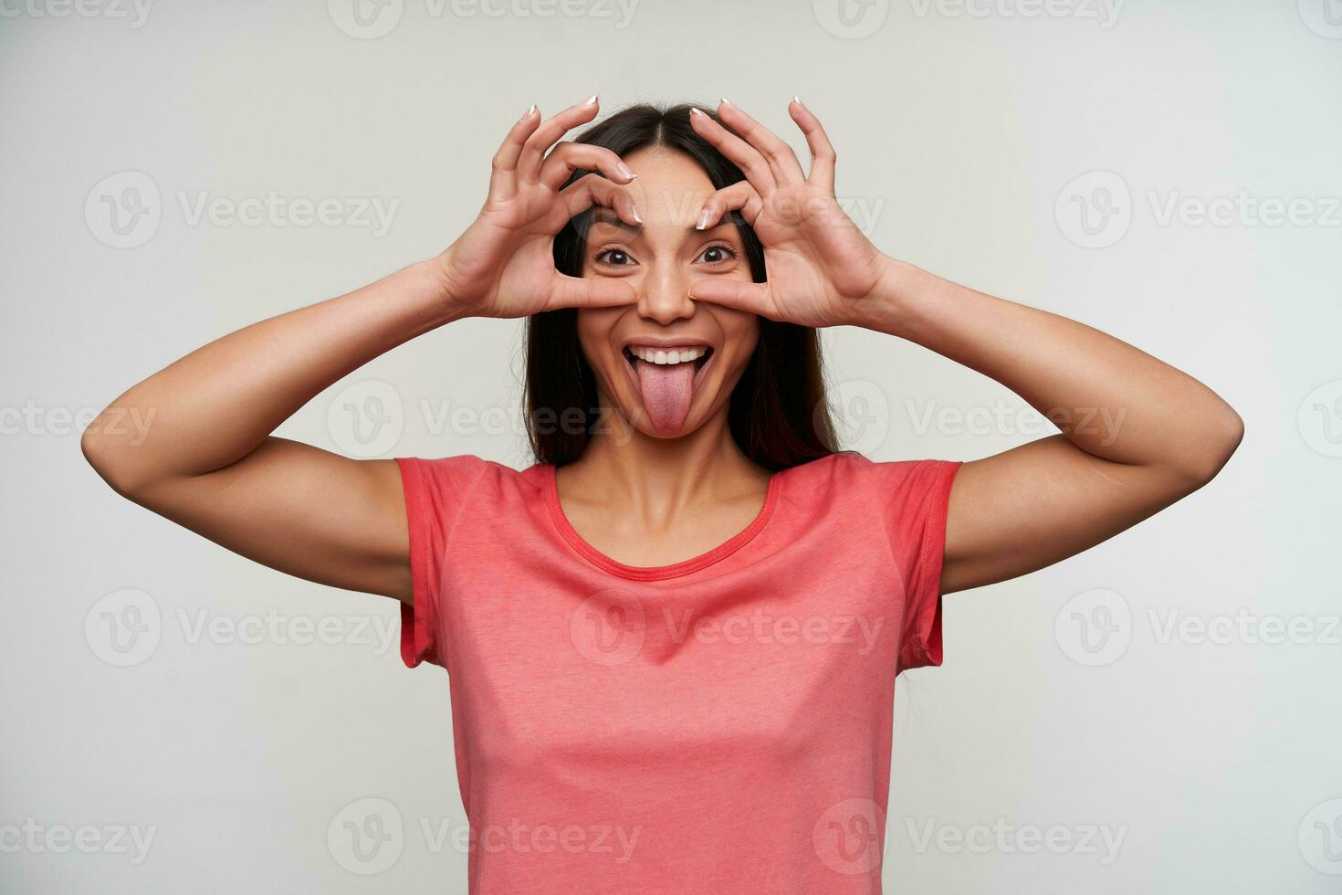 Funny young dark haired joyful woman in pink t-shirt folding eyewear from raised hands while looking cheerfully to camera and showing her tongue, isolated over white background photo