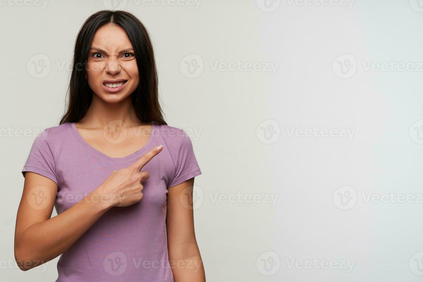 Studio photo of young brown-eyed dark haired lady showing aside with index finger and grimacing discontentedly her face while looking at camera, isolated over white background