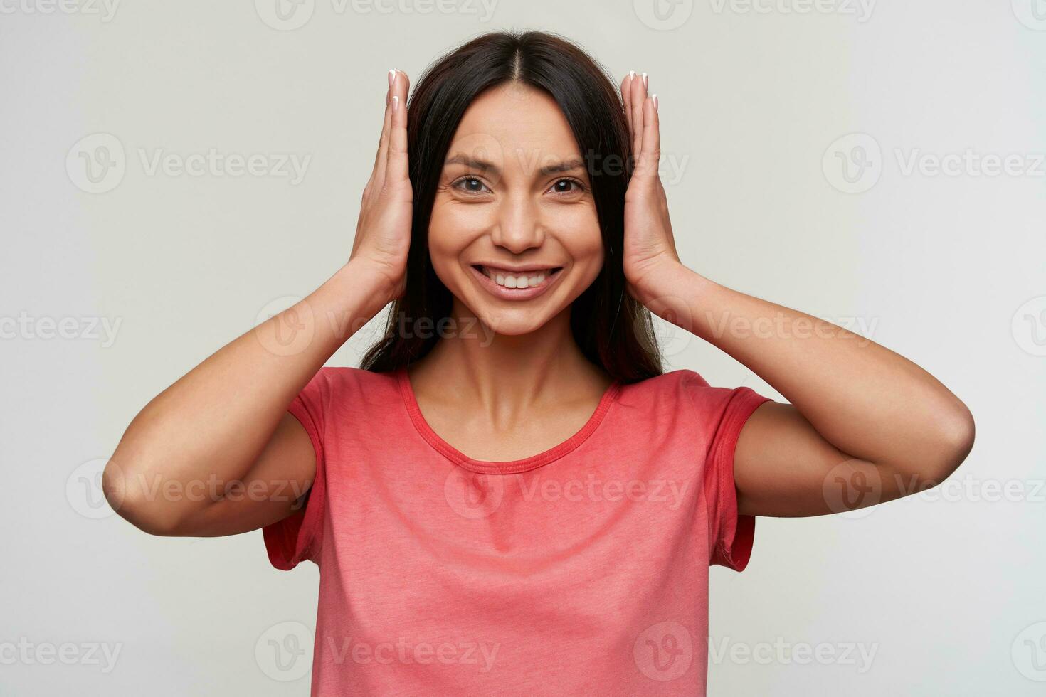 Cheerful young lovely dark haired lady with natural makeup closing her ears with hands and looking happily at camera and smiling widely, standing against white background photo