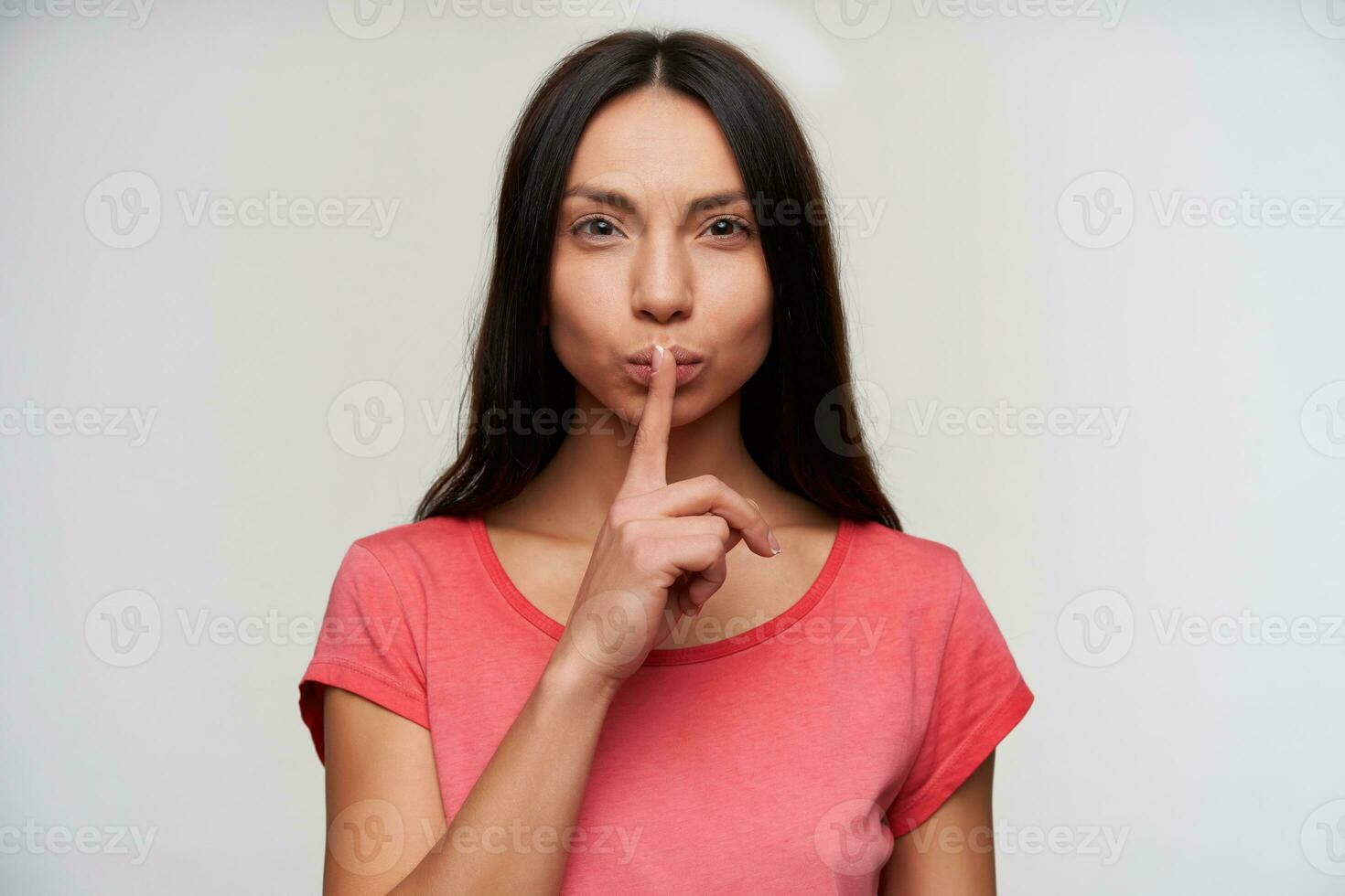 Portrait of charming young brunette woman raising forefinger in hush gesture and looking at camera with serious face, asking to be quiet while standing over white background photo