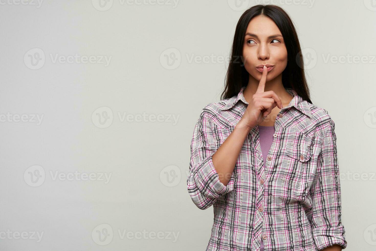 Thoughtful young lovely brunette woman with casual hairstyle looking pensively aside and keeping forefinger on her lips, dressed in purple t-shirt and checkered shirt over white background photo