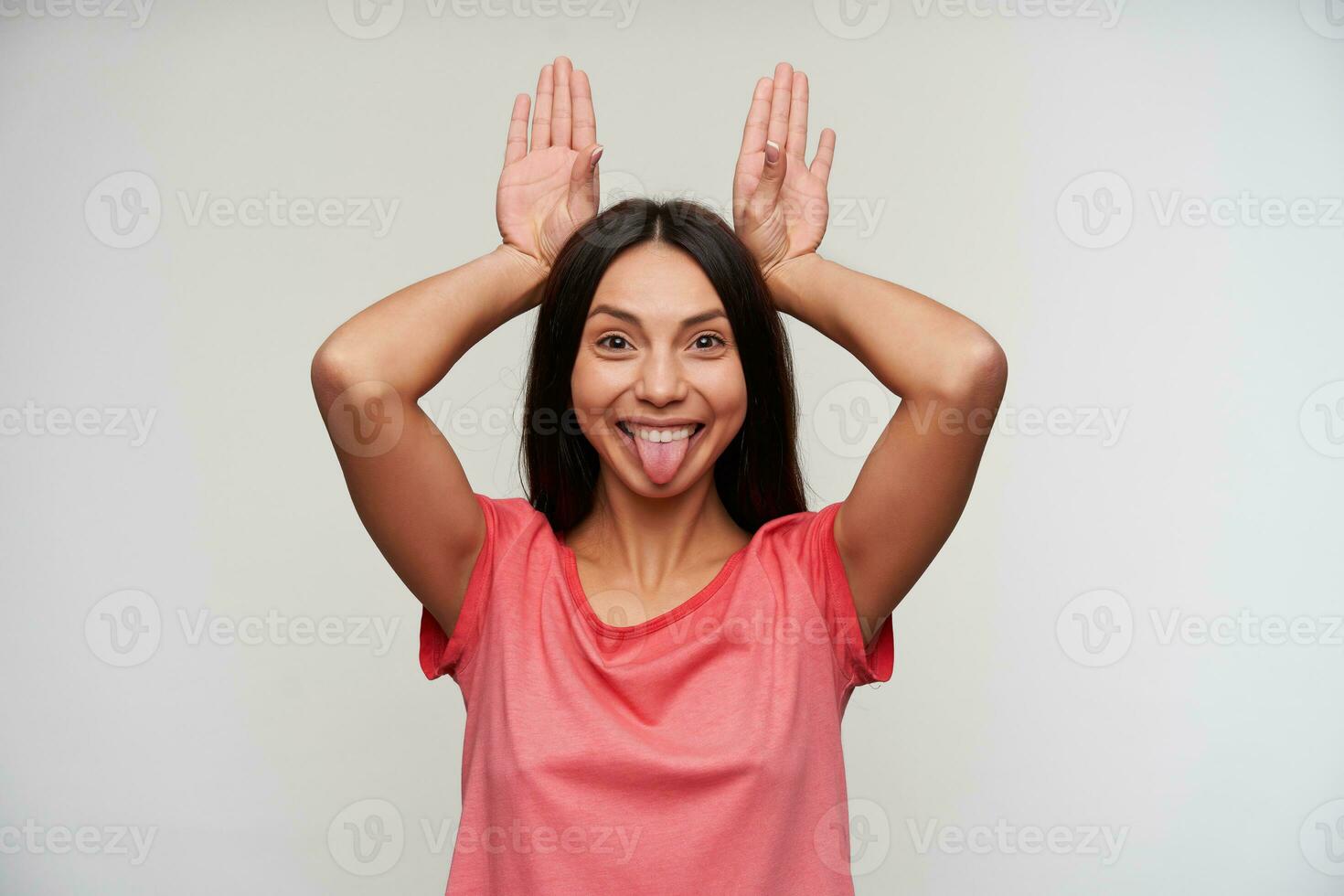 Funny photo of pretty young brown-eyed brunette female imitating rabbit ears with raised hands and showing happily her tongue to camera, posing over white background