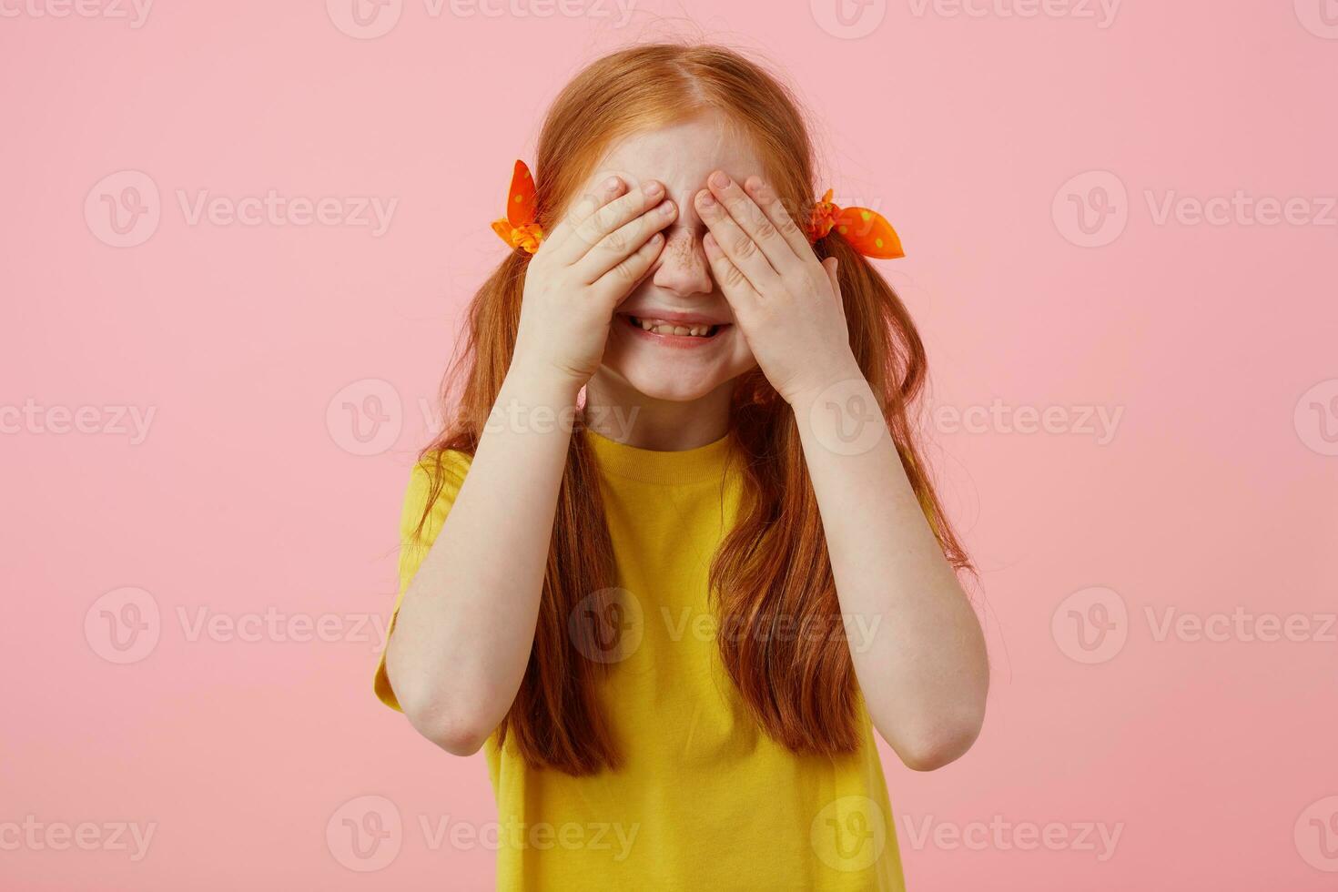 Photo of little freckles red-haired girl with two tails, smiles and covers eyes with palms, wears in yellow t-shirt, stands over pink background.