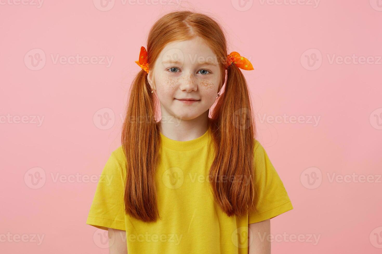 retrato pequeño pecas Pelirrojo niña con dos cruz, mira dentro el cámara y sonrisas, usa en amarillo camiseta, soportes terminado rosado antecedentes. foto