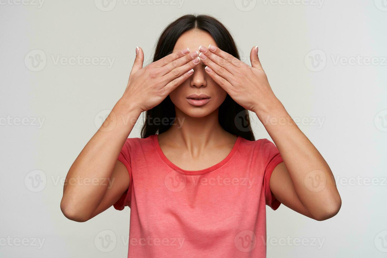 Good looking young brunette female with natural makeup holding raised palms on her eyes while posing over white background, wearing casual pink t-shirt photo