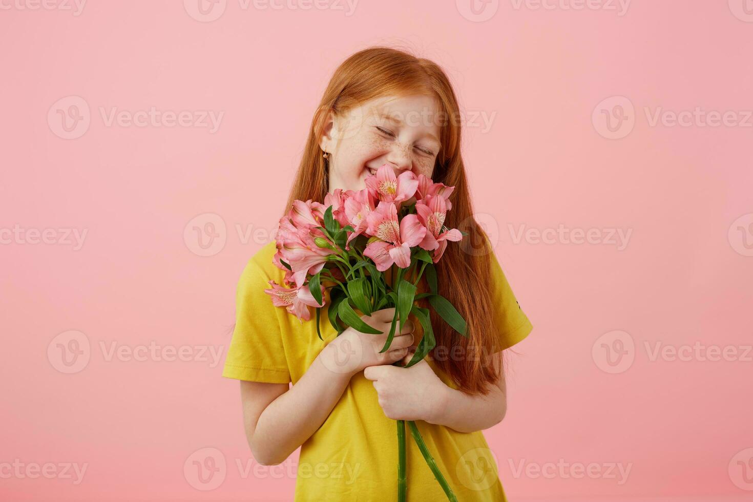 Portrait petite smiles freckles red-haired girl with two tails, looks cute, wears in yellow t-shirt, holds bouquet and stands over pink background, enjoying the smell of flowers. photo