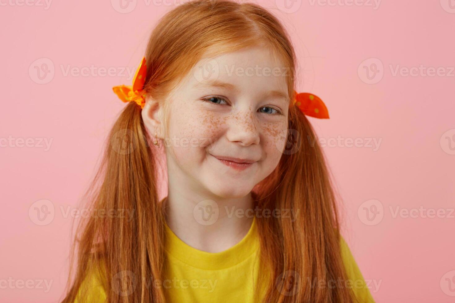 cerca arriba de sonriente chiquita pecas Pelirrojo niña con dos cruz, sonriente y mira lindo, usa en amarillo camiseta, soportes terminado rosado antecedentes. foto