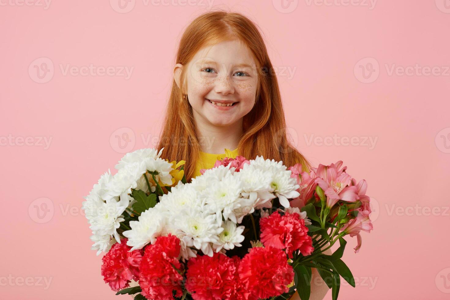 Happy petite freckles red-haired girl with two tails, broadly smiling and looks cute, wears in yellow t-shirt, holds bouquet and stands over pink background. photo