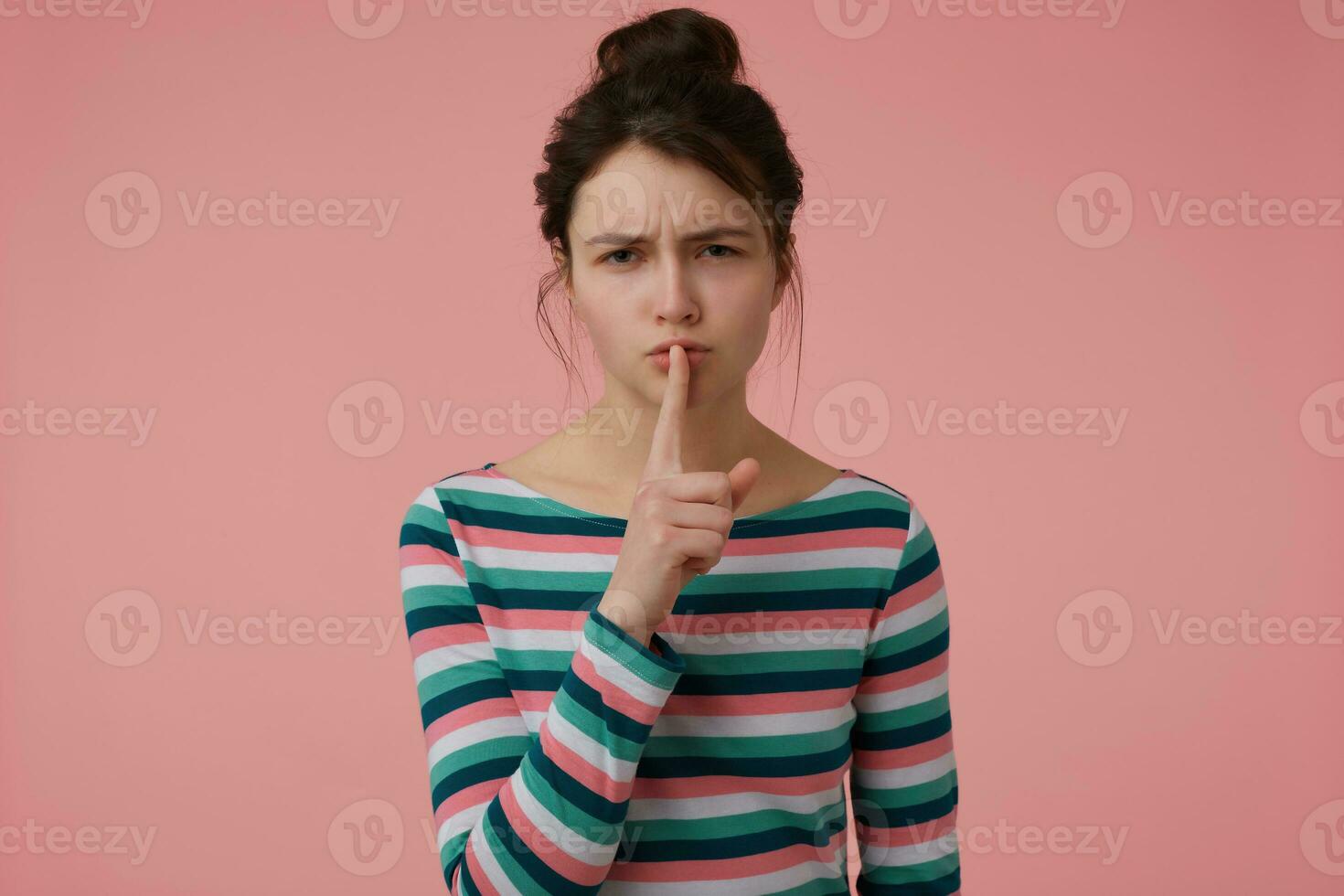 joven dama, bonito mujer con morena pelo y bollo. vistiendo a rayas blusa y demostración firmar de silencio, conmovedor labios con un dedo. acecho a el cámara aislado terminado pastel rosado antecedentes foto