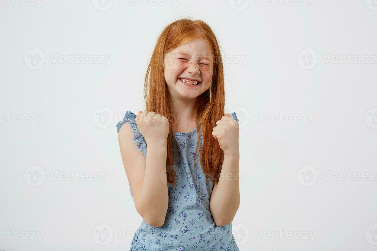 Portrait of happy smiling petite freckles red-haired girl, lwears in bluet, stands over white background with fists up. photo