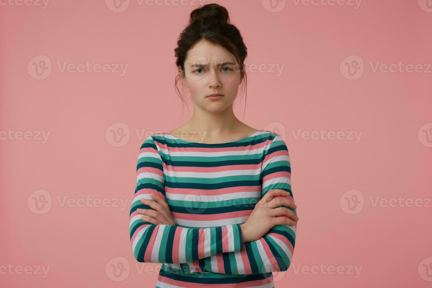 Young lady, angry, serious looking woman with brunette hair and bun. Wearing striped blouse and fold hands on a chest. Emotional concept. Watching at the camera isolated over pastel pink background photo