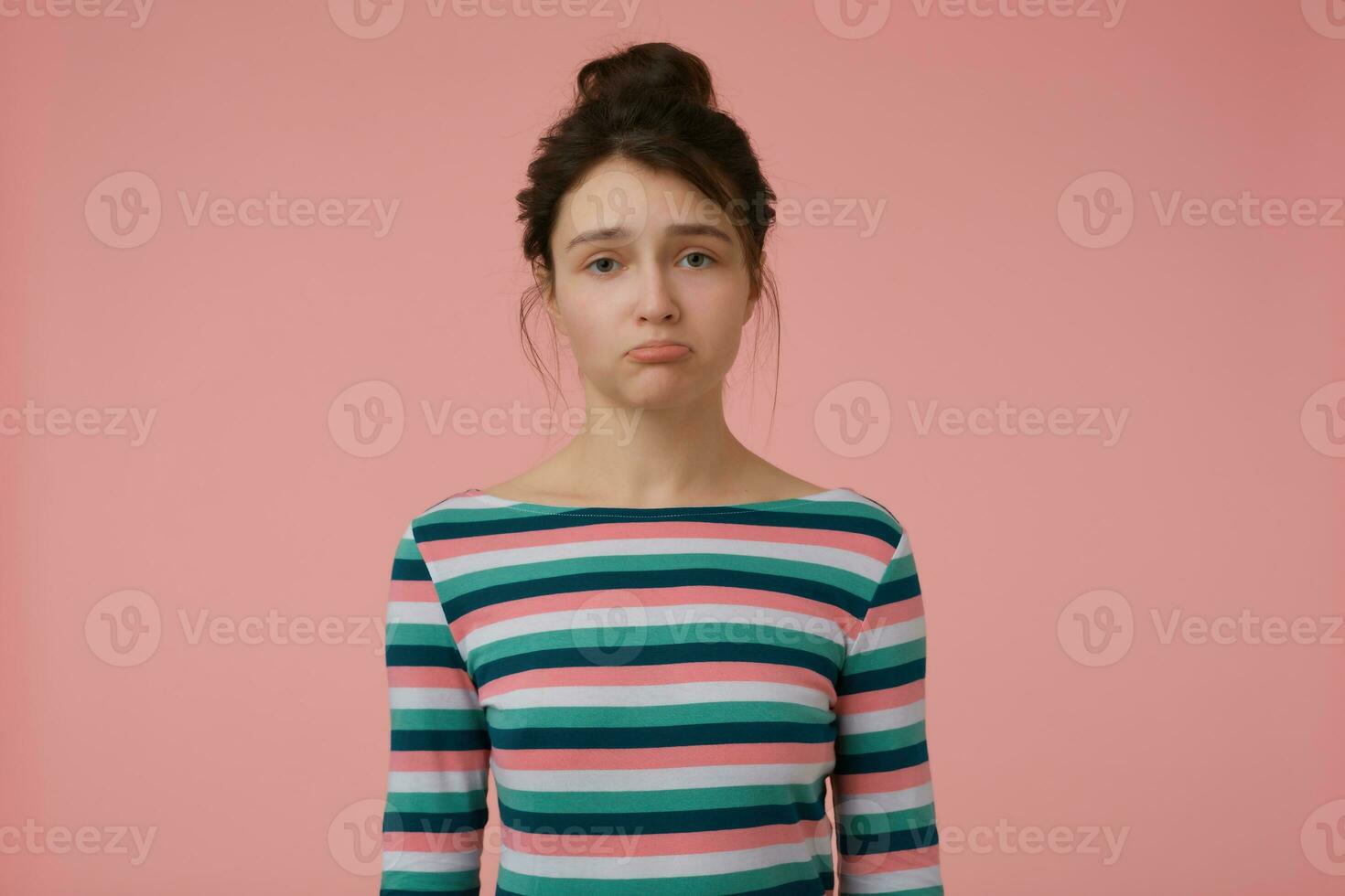 joven dama, infeliz mujer con morena pelo y bollo. vistiendo a rayas blusa y abadejo un labio, ofendido emocional concepto. acecho a el cámara aislado terminado pastel rosado antecedentes foto