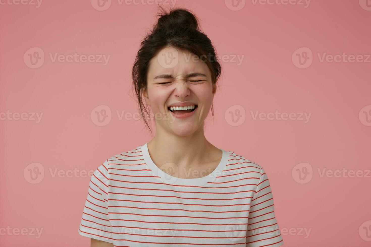 Portrait of attractive, joyful girl with brunette hair and bun. Wearing t-shirt with red strips and laughing with closed eyes. Emotional concept. Stand isolated over pastel pink background photo