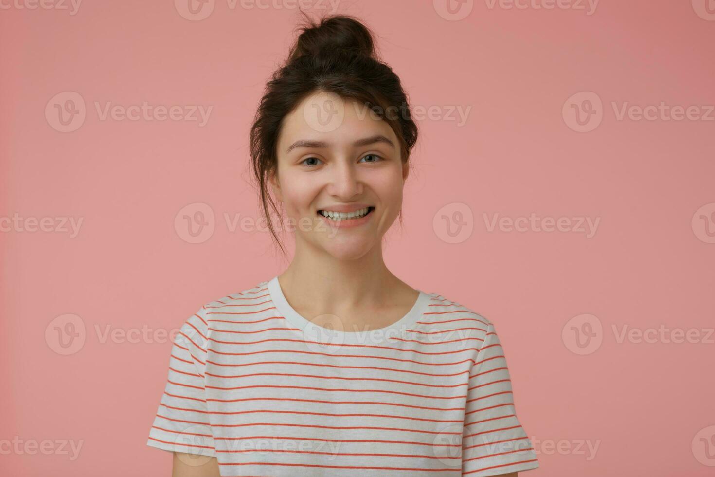 Teenage girl, happy looking woman with brunette hair and bun. Wearing t-shirt with red strips and big smile. Emotional concept. Watching at the camera isolated over pastel pink background photo