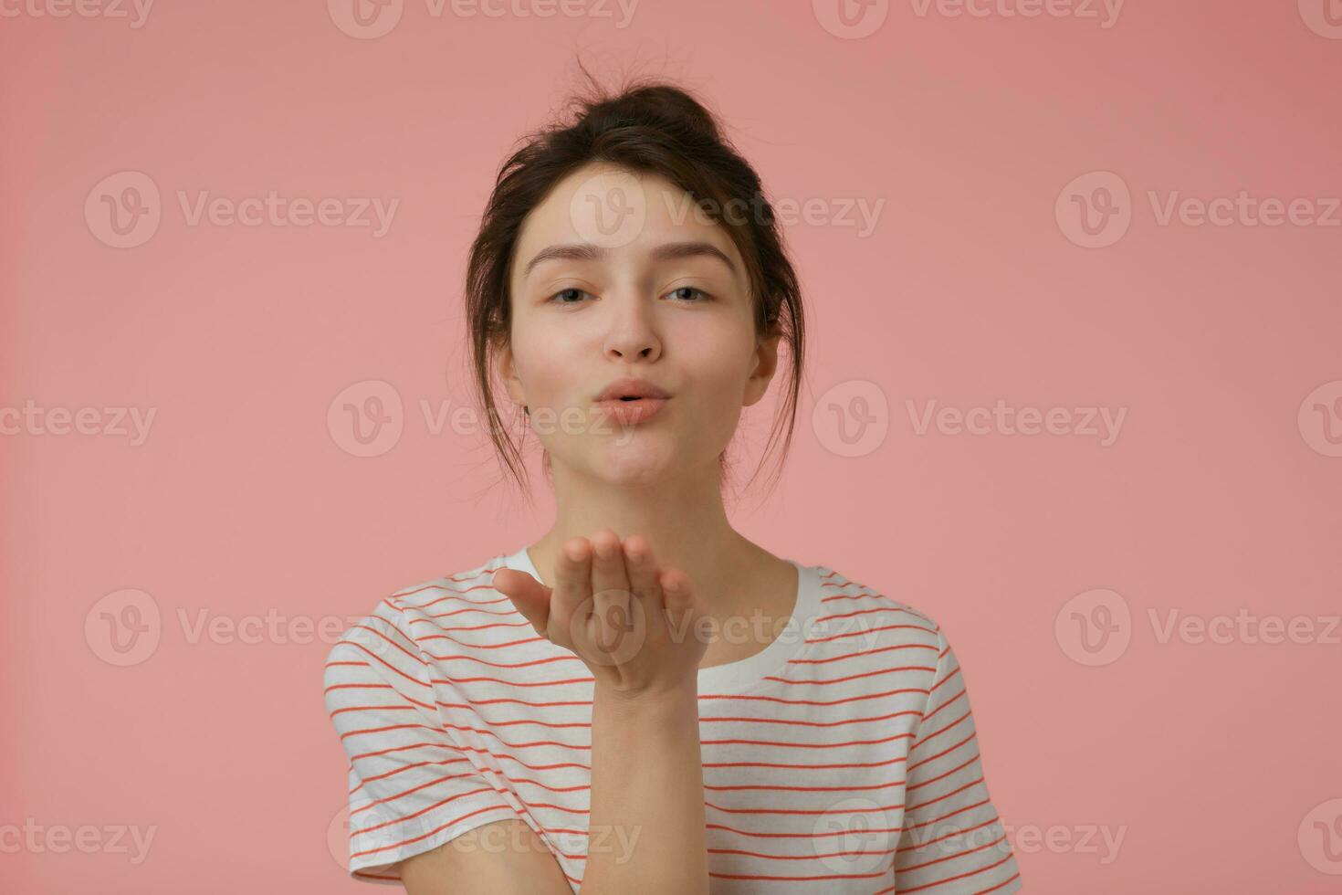 joven dama, bonito mujer con morena pelo y bollo. vistiendo camiseta con rojo tiras y enviando un beso. emocional concepto. acecho a el cámara aislado terminado pastel rosado antecedentes foto