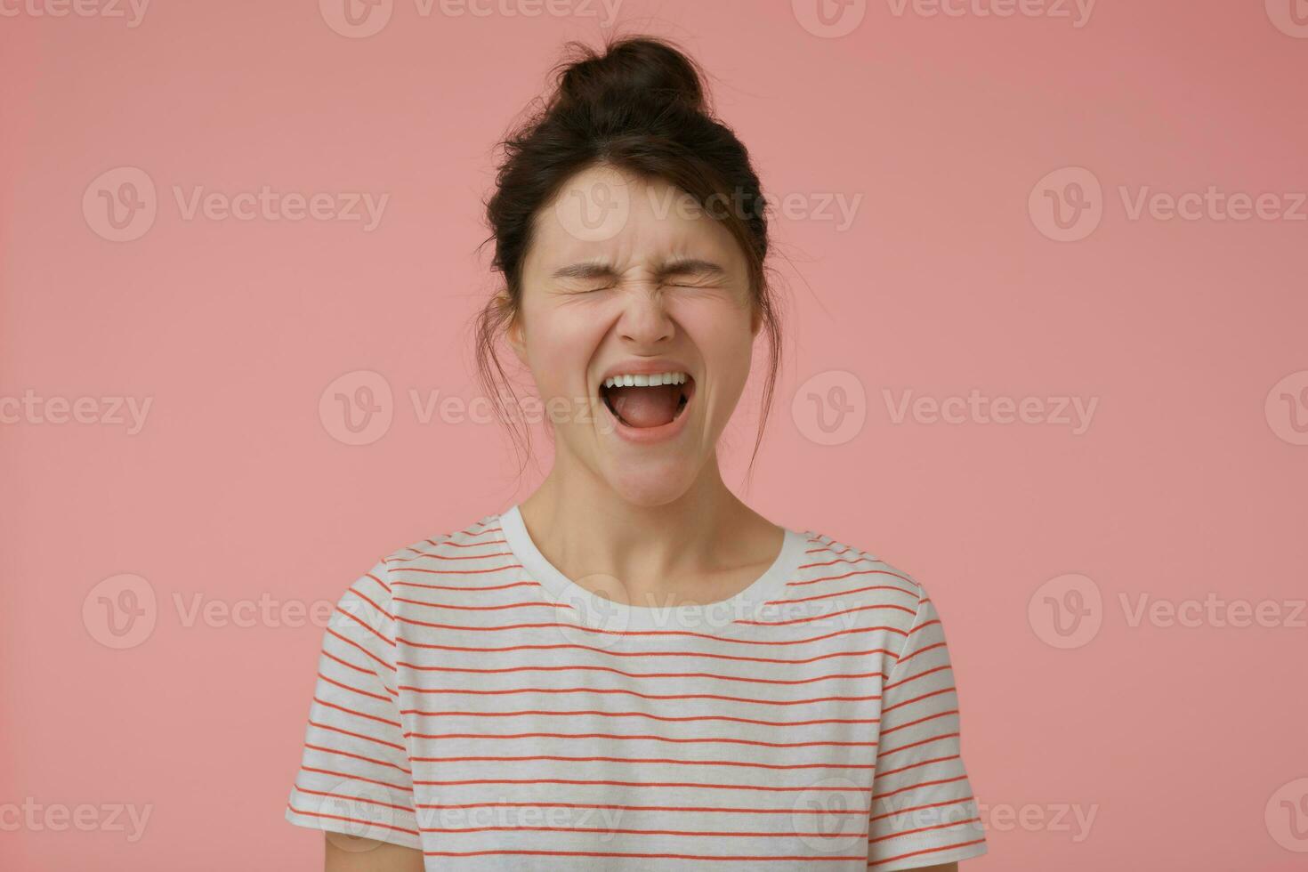 Teenage girl, angry looking woman with brunette hair and bun. Wearing t-shirt with red strips and yelling in anger. Emotional concept. Stand isolated over pastel pink background photo