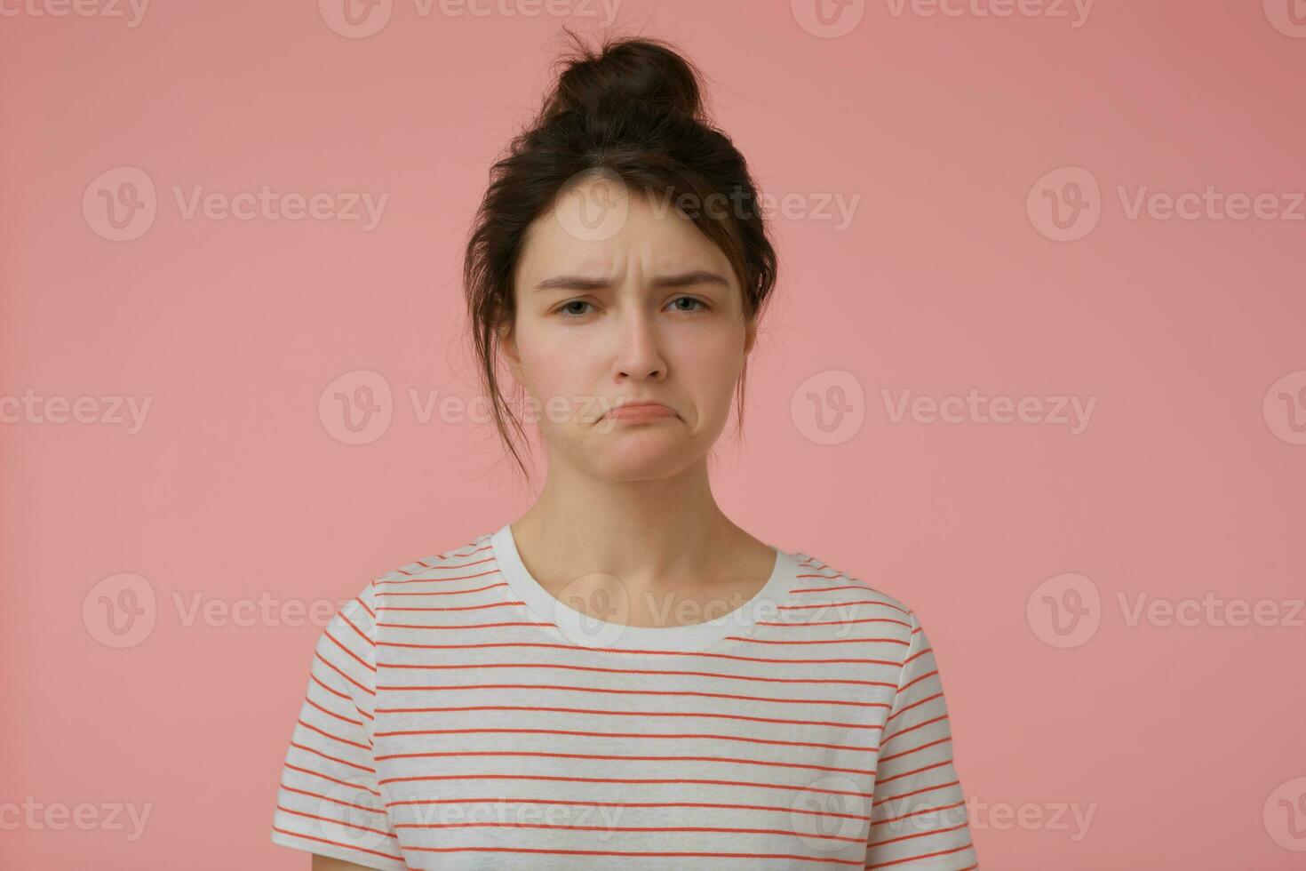 Young lady, unsatisfied woman with brunette hair and bun. Wearing t-shirt with red strips and looking offended. Emotional concept. Watching at the camera isolated over pastel pink background photo