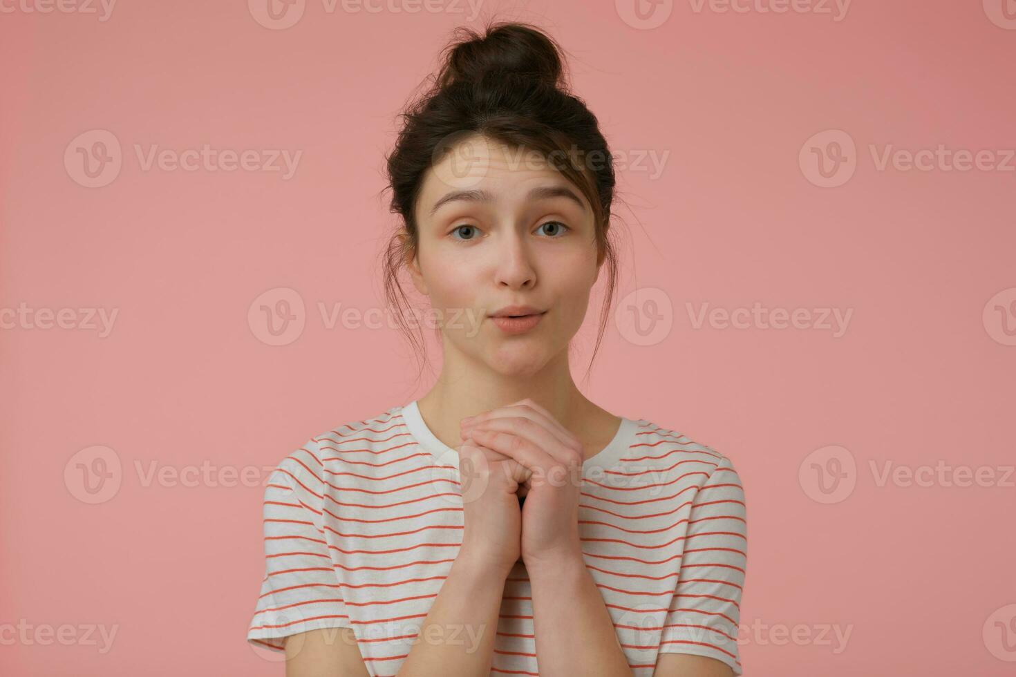 Portrait of attractive, brunette girl with brunette hair and bun. Wearing t-shirt with red strips and begging you. Emotional concept. Watching at the camera isolated over pastel pink background photo