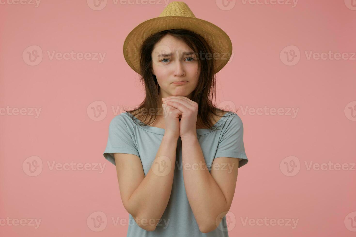 Teenage girl, woman with begging look and long brunette hair. Wearing blueish t-shirt and hat. Holding hands under her chin and ask. Watching at the camera isolated over pastel pink background photo