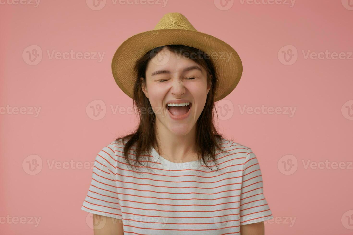 Teenage girl, happy looking woman with long brunette hair. Wearing t-shirt with red strips and hat. Emotional concept. Screams with closed eyes. Stand isolated over pastel pink background photo