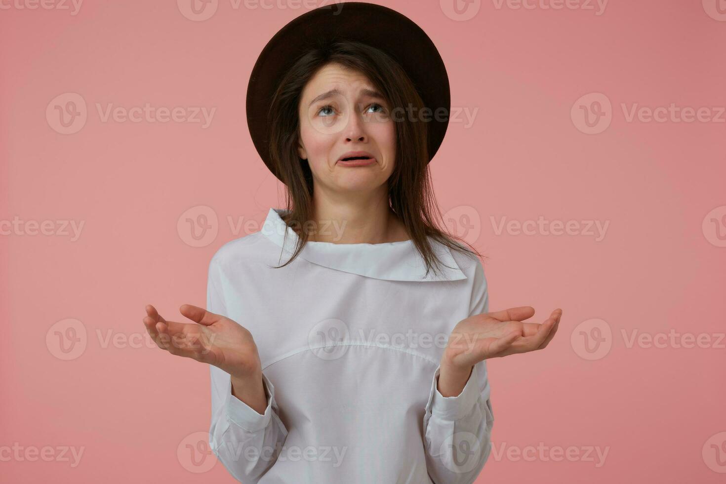 Upset looking woman, unhappy girl with long brunette hair. Wearing white blouse and black hat. Emotional concept. Stand isolated over pastel pink background and watching up at copy space photo